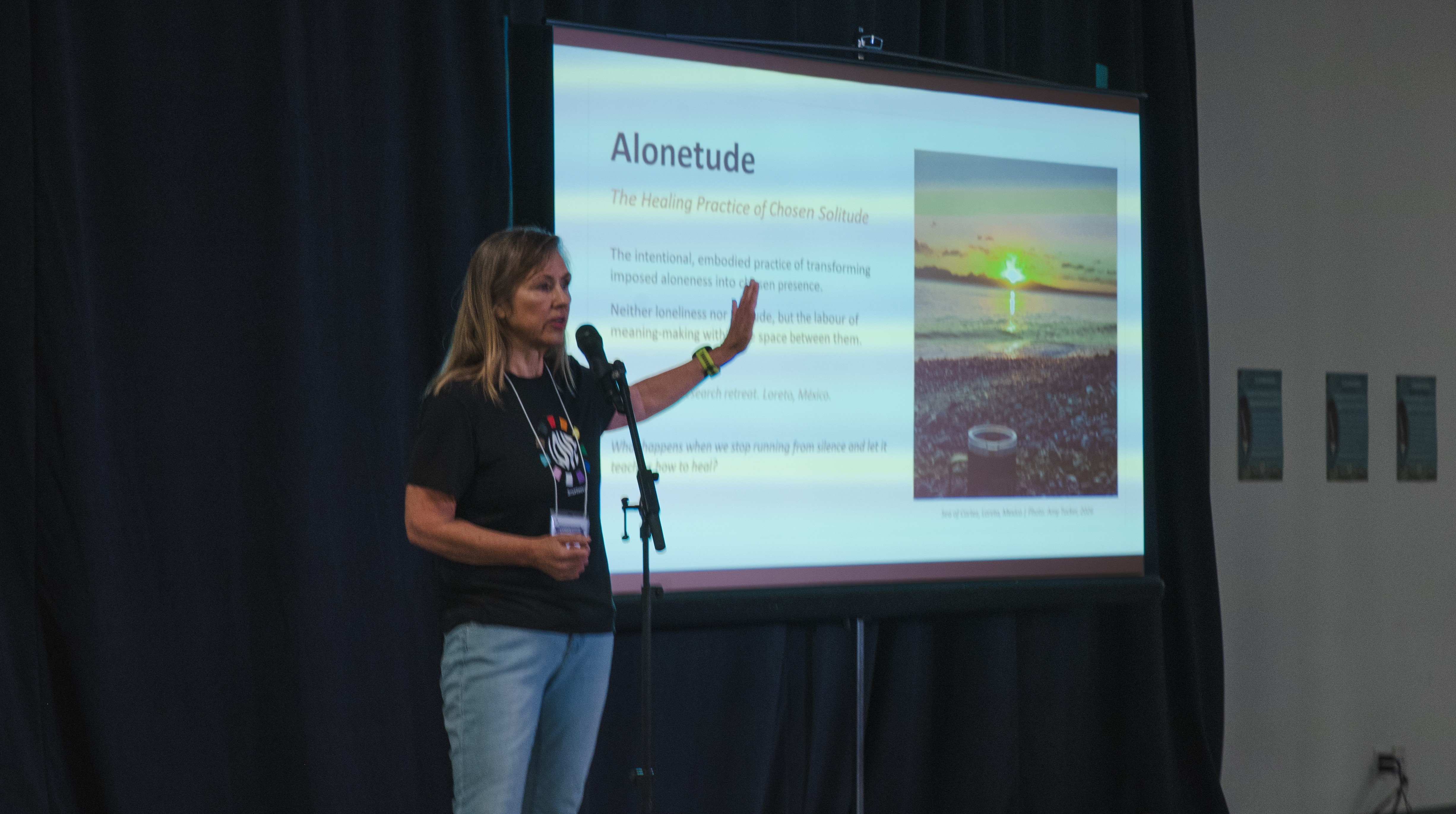 Amy Tucker presenting at the 3 Minute Thesis competition at Thompson Rivers University, standing at a podium with a slide showing the word Alonetude behind her