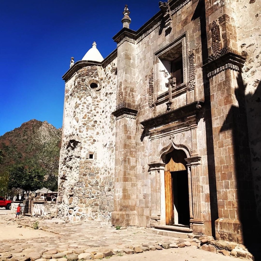 The stone facade and carved wooden doorway of the Misión San Francisco Javier de Viggé-Biaundó near Loreto, Baja California Sur, with rough rubble walls on the left and ornate stonework framing the entrance, desert mountains rising behind, January 2026