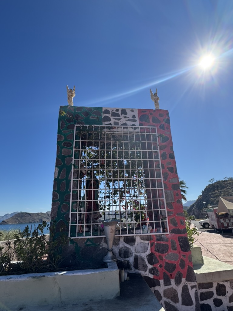 A roadside shrine in Loreto painted in the colours of the Mexican flag with two angel figures on top, the sea and mountains visible behind