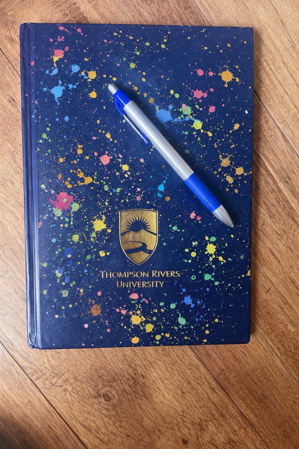 A Thompson Rivers University branded journal with a colourful paint-splattered cover, a pen resting on top, on a wooden surface
