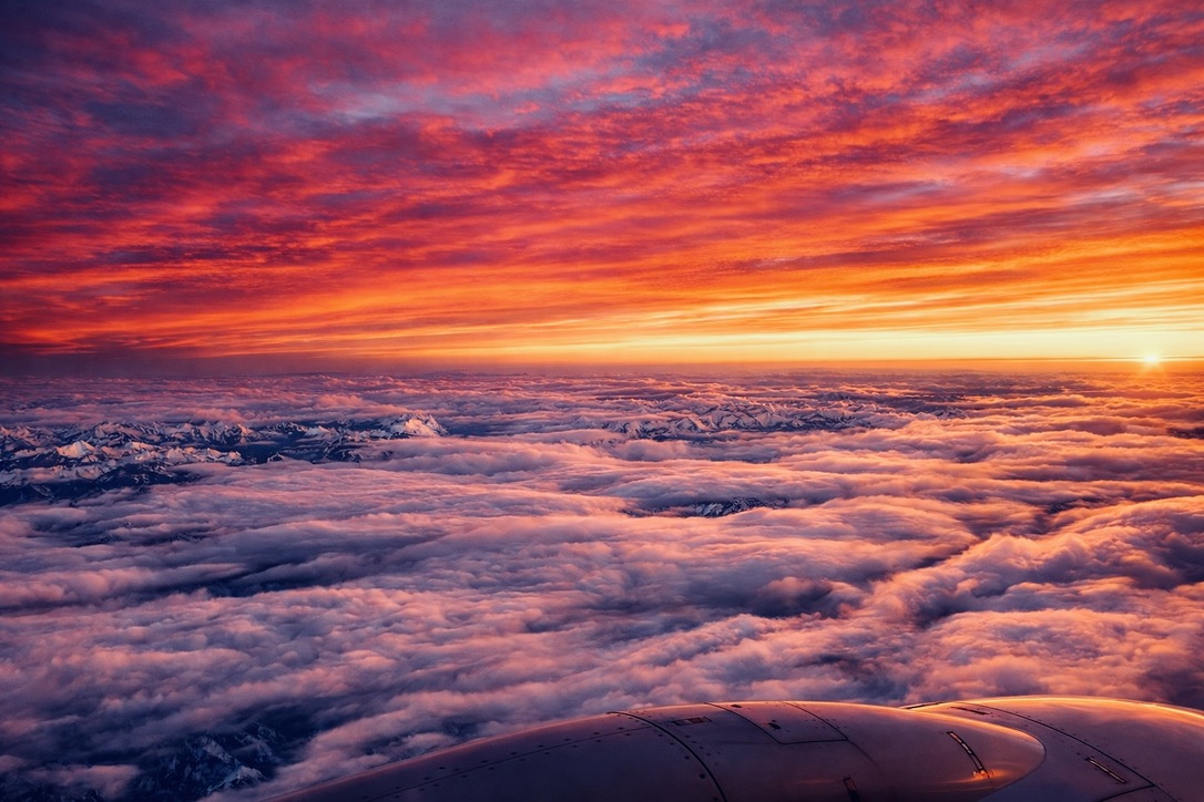 Aerial view of a blazing sunset above a sea of clouds, with snow-capped mountain peaks visible below, photographed from an airplane window.