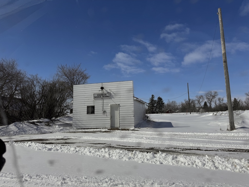 A small isolated white building labelled Insinger Water Works standing alone in a vast snow-covered prairie landscape under a bright blue winter sky.