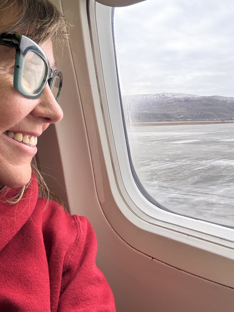 A woman in a red sweater smiling while looking out an airplane window at a frozen, pale landscape below a white sky.