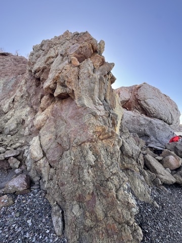 A large weathered rock formation standing at the shore under an open sky, its surface layered and worn by time.