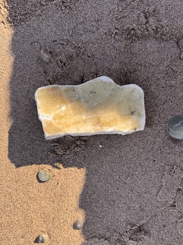 A pale flat stone with golden and cream tones resting in dark sand, smooth-edged and quietly present.